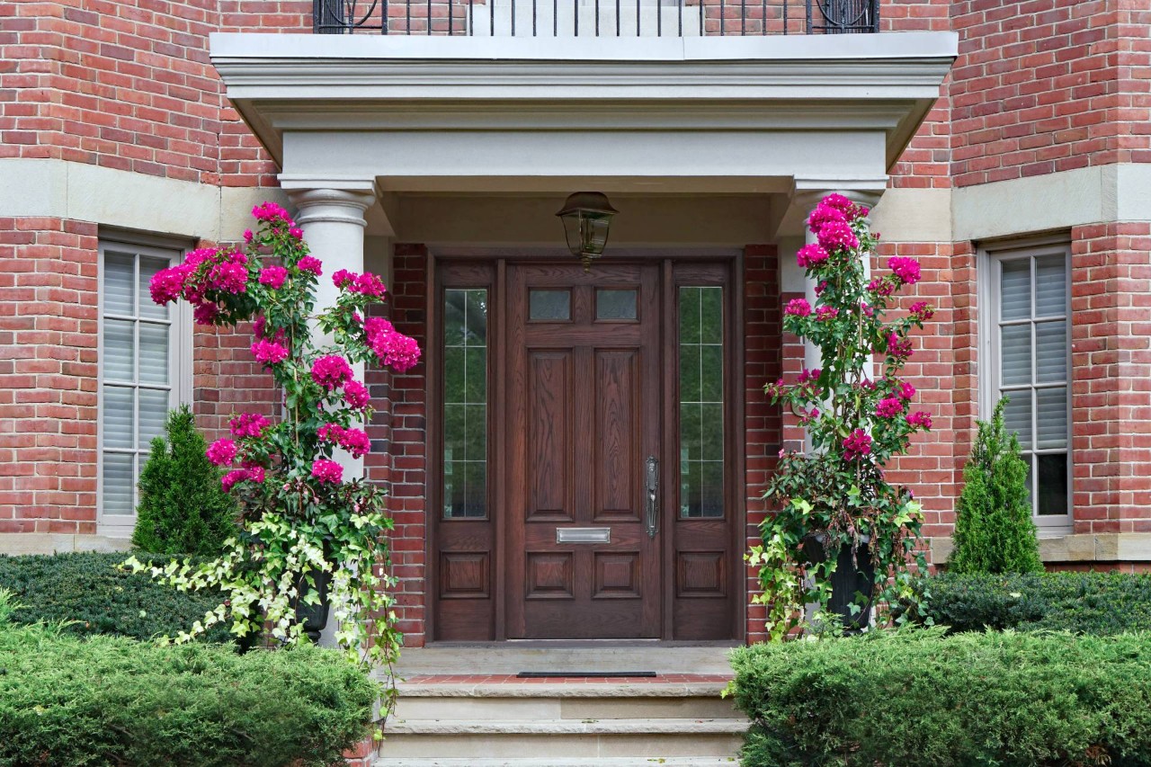 Picturesque brick home with pink flowers, manicured greenery, and a dark wood front door with sidelights at Window Accents & Flooring near Princeton New Jersey (NJ)